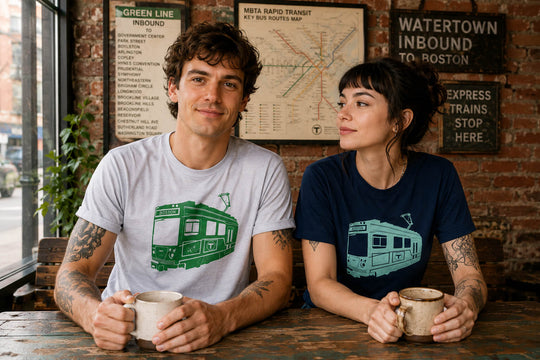 A young man and woman in coffee shop wearing silver and navy blue Boston MBTA green line trolley t-shirts
