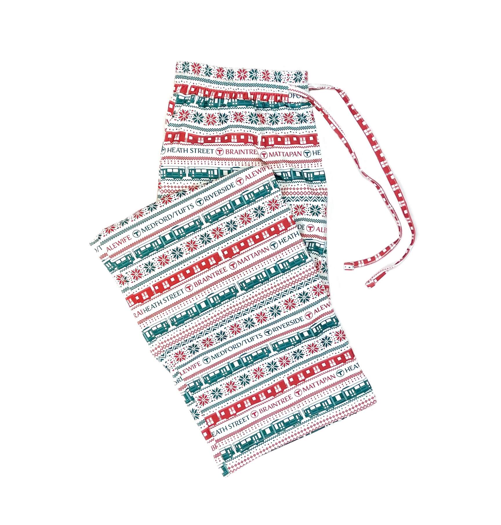 A folded pair of adult-size ivory pajama bottoms with a green and red pattern of MBTA trolleys and trains, snowflakes, and destination station names