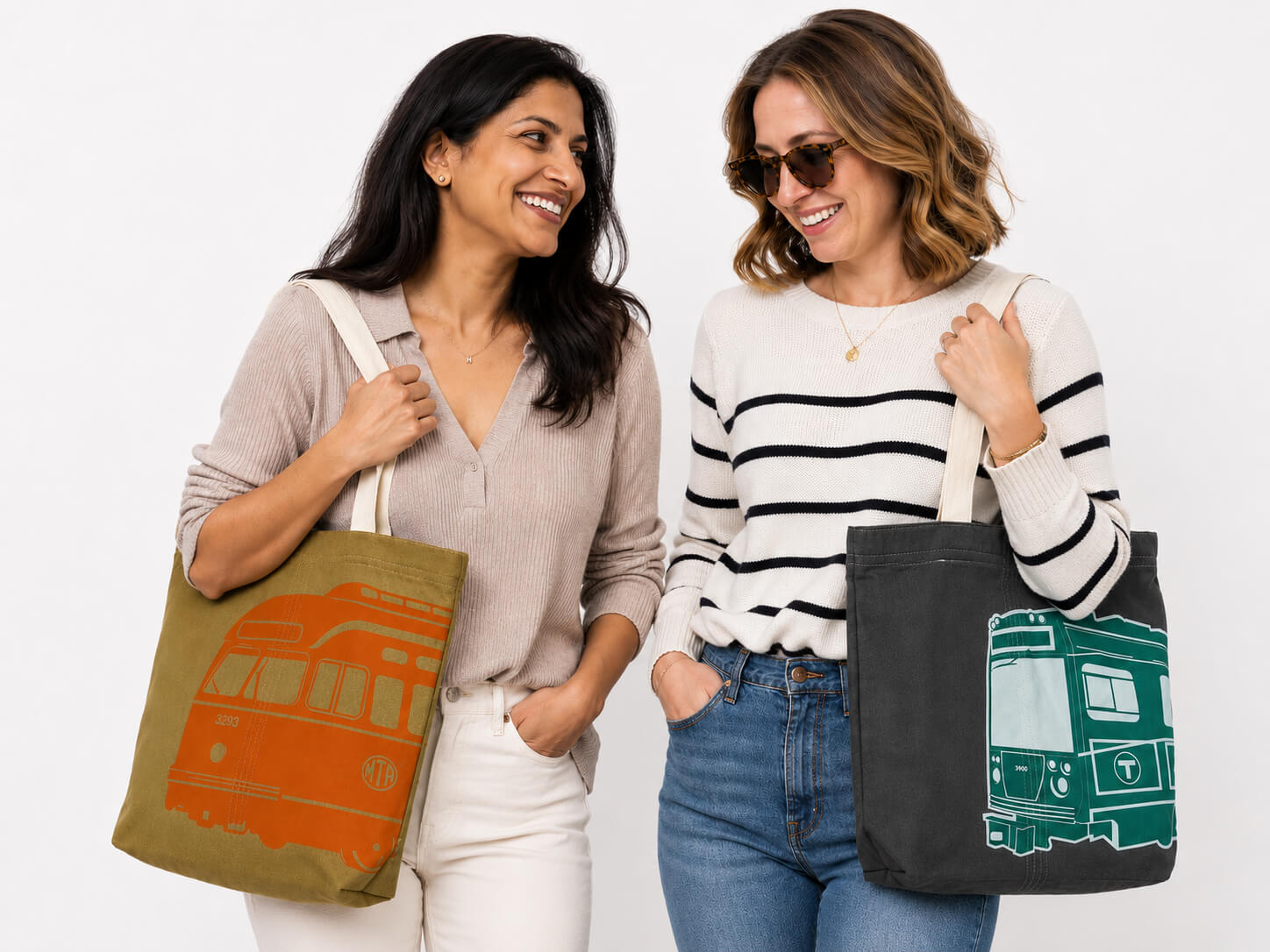 Two women with over-the-shoulder fashion Boston MBTA tote bags - one with the PCC Bus wheat-colored bag and the other with the grey Green Line Trolley bag