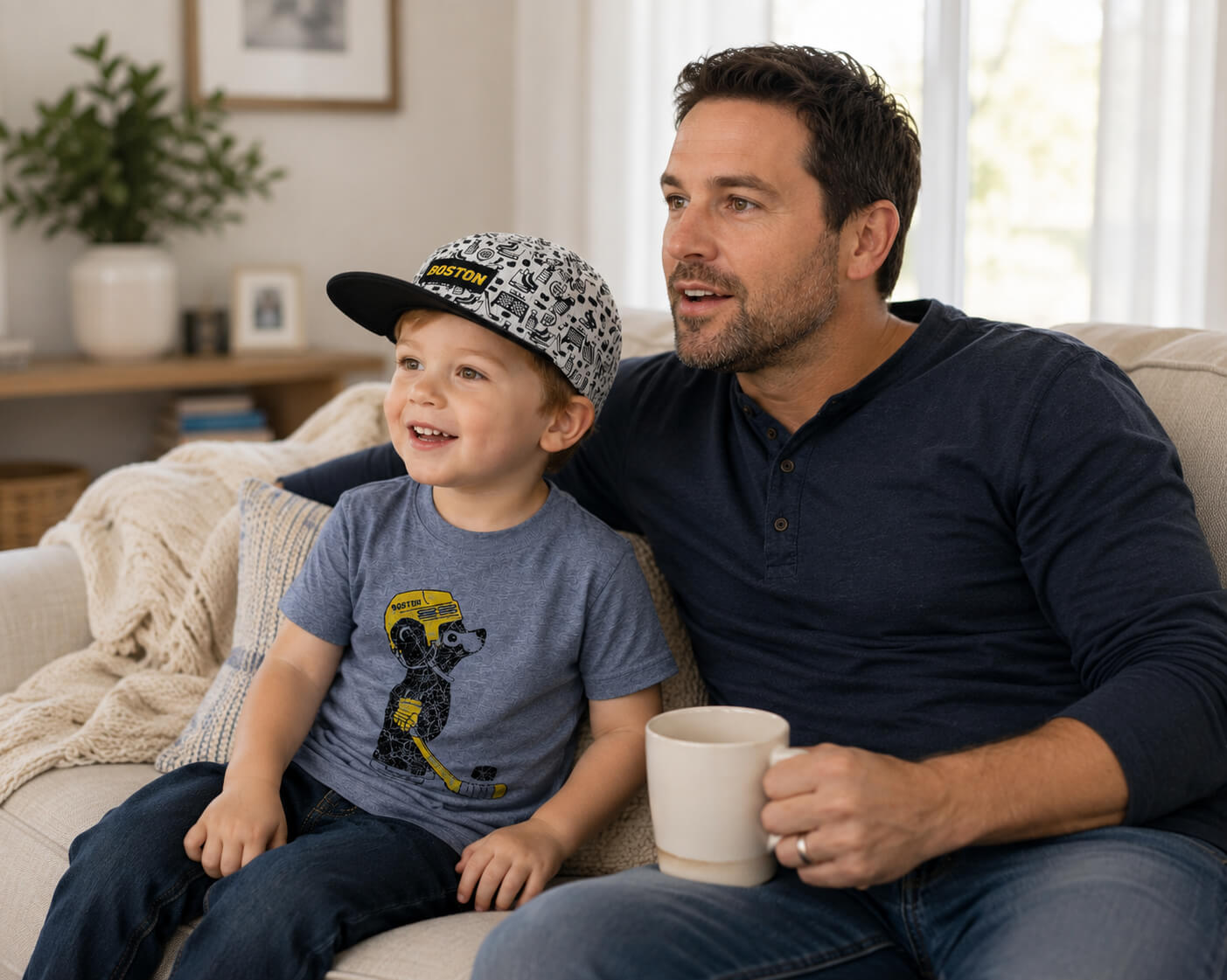 A father and son sitting together on a beige couch in a bright living room. The son is wearing a cute Boston Hockey cub t-shirt and a and a grey, black and gold hockey-patterned baseball cap.