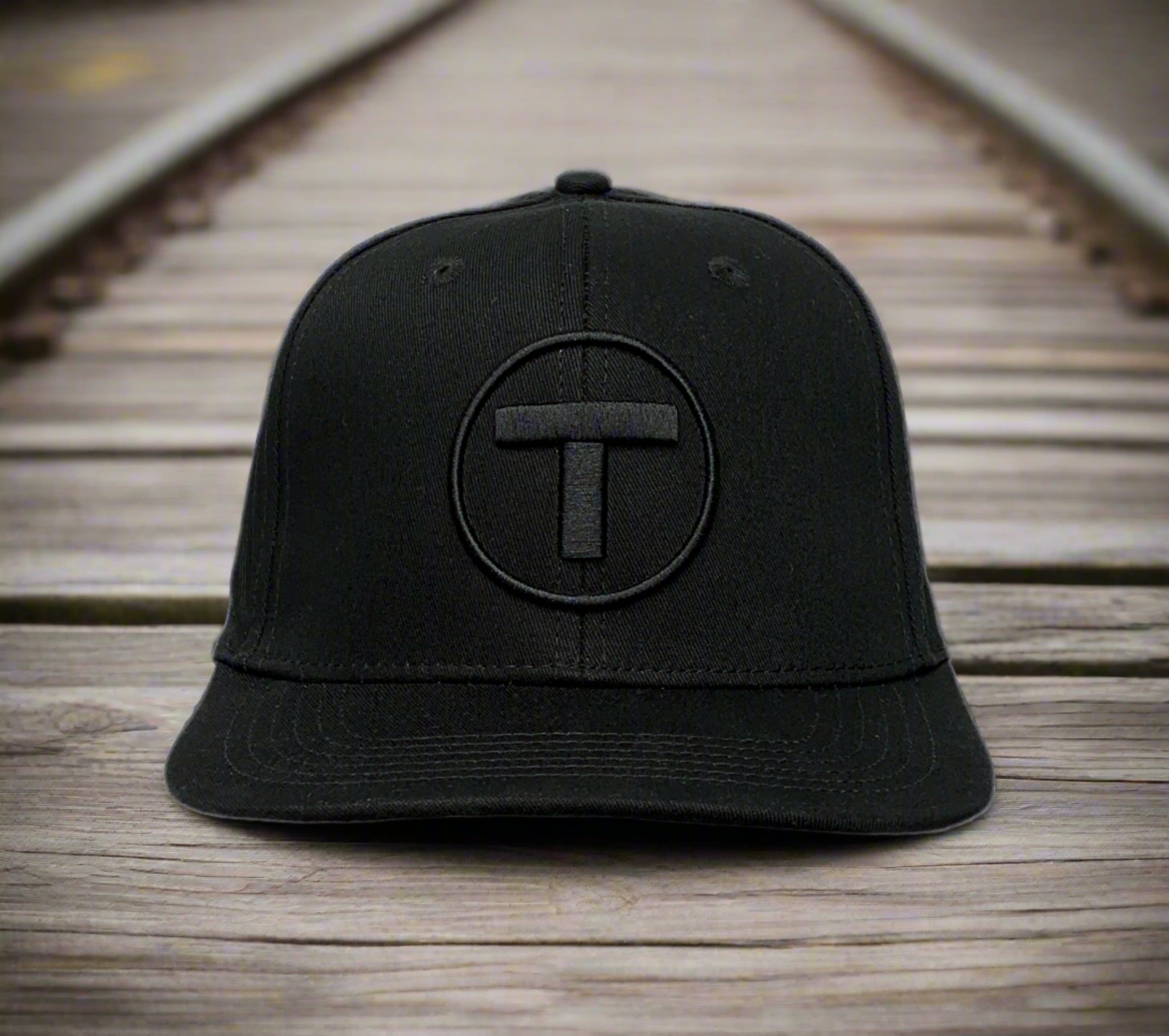 Front view of black flat brim adult cap with puff embroidered MBTA T Logo